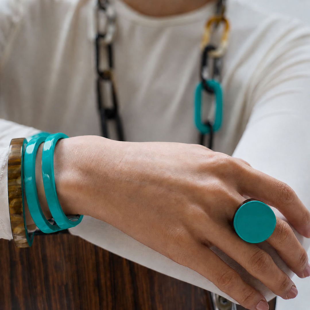 Close-up of a hand wearing a Branch Jewellery round turquoise horn ring and bracelets on a neutral background. 