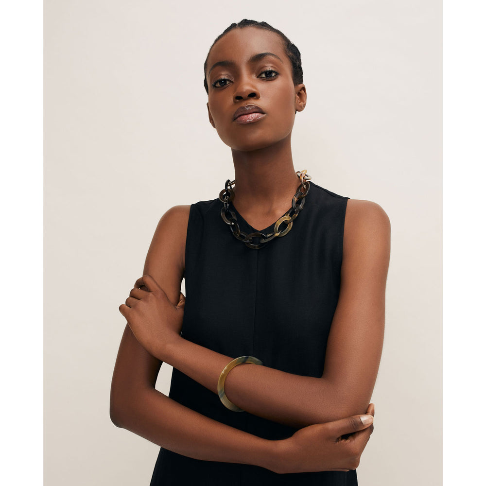 A person in a sleeveless black dress stands confidently against a neutral background, arms crossed. Their look is elevated by Branch Jewellery’s chunky handcrafted buffalo horn necklace and Brown Natural flat horn Bangle, ethically sourced and handmade in Vietnam.