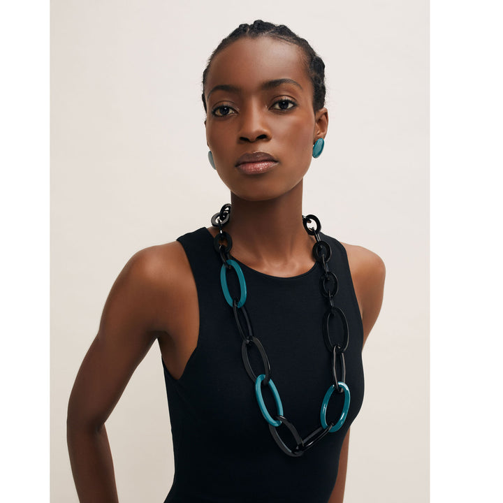 A person is posed against a plain background wearing a sleeveless black top, an expertly handmade Branch Jewellery Oval Link Black & Blue Horn Necklace, and matching earrings. Crafted in Vietnam, the jewelry features striking black and turquoise buffalo horn links.