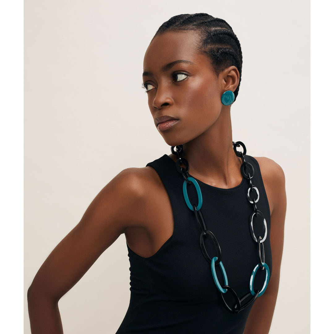 A woman with short braided hair wears a sleeveless black top, blue stud earrings, and the Oval Link Black & Blue Horn Necklace by Branch Jewellery. She looks to her left against a plain background.