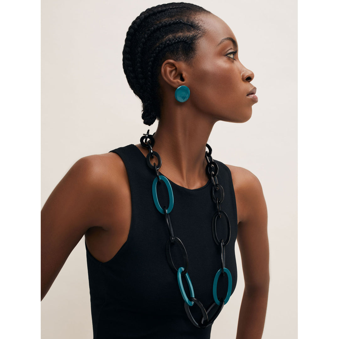 A person with braided hair and wearing a black sleeveless top, large teal earrings, and the standout Branch Jewellery Oval Link Black & Blue Horn Link Necklace, handmade in Vietnam, is facing right.