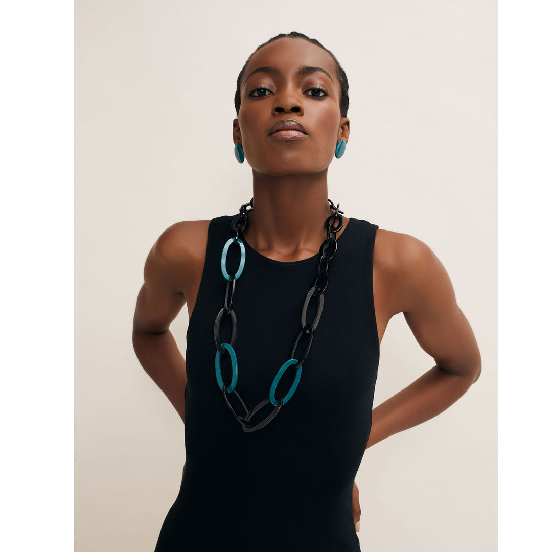 A person in a sleeveless black dress accessorizes with the Branch Jewellery Oval Link Black & Blue Horn Necklace and matching blue earrings, ethically sourced and handmade in Vietnam, set against a plain background.