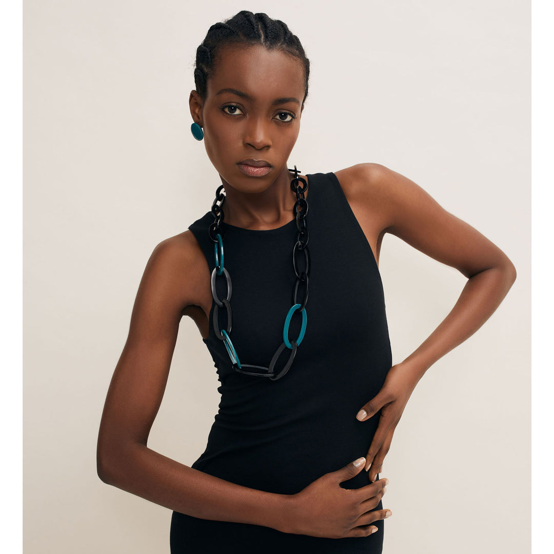 A person wears a sleeveless black dress with Branch Jewellerys Oval Link Black & Blue Horn Necklace, complemented by matching teal earrings. This unique, handmade piece from Vietnam enhances the ensemble with ethically sourced buffalo horn in black and teal.